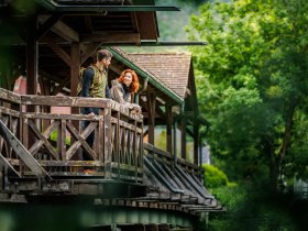 Ein Paar steht auf einer &uuml;berdachten Holzbr&uuml;cke und schaut sich an, umgeben von gr&uuml;ner Natur.
