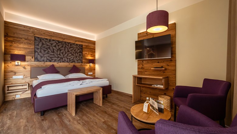 Gemütliches Hotelzimmer mit Holzwänden, Doppelbett, TV und lila Sesseln.