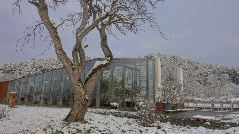 Eine moderne Orangerie im Winter, umgeben von schneebedeckten B&auml;umen und H&uuml;geln.