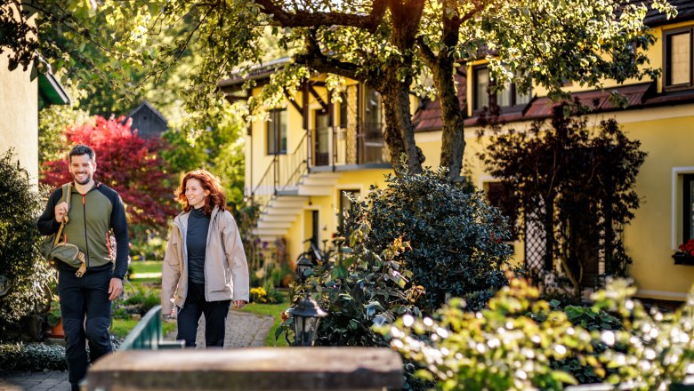 Ein Mann und eine Frau in Wanderkleidung gehen durch den grünen, gepflegten Garten der Pension Hendling