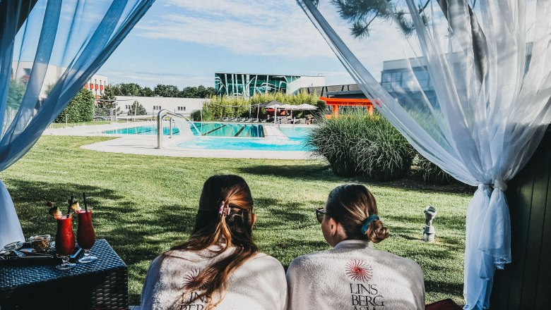 Zwei Personen in Bademänteln liegen in einer Cabana mit Blick auf einen Pool in der Therme Linsberg Asia.
