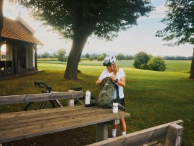 Eine Frau mit Fahrradhelm steht an einem Picknicktisch in einer l&auml;ndlichen Umgebung und packt ihren Rucksack. Im Hintergrund ist eine kleine Kapelle zu sehen.