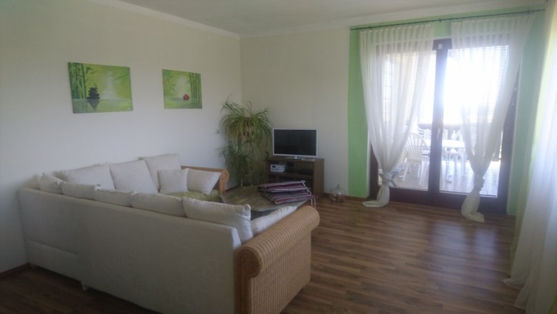 Ein Wohnzimmer mit einem weißen Sofa, einem kleinen Fernseher, Pflanzen und großen Fenstern mit Vorhängen.