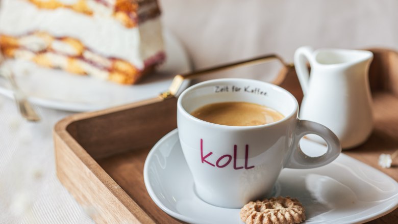 Eine Tasse Kaffee mit einem Keks auf einem Tablett, im Hintergrund ein Stück Torte.