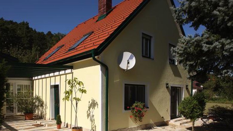 Gelbes Haus mit rotem Dach und Satellitenschüssel, umgeben von Bäumen.