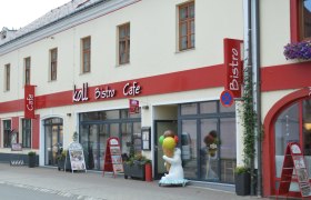Au&szlig;enansicht des Koll Bistro Caf&eacute;s mit rotem Schild und Eisb&auml;rfigur mit Eis-Stanizel vor dem Eingang.