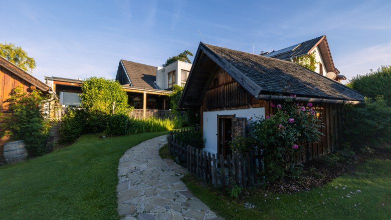 Ein kleiner Gartenweg führt zu einem traditionellen Holzhaus mit Blumen und Büschen, umgeben von weiteren Gebäuden unter blauem Himmel.