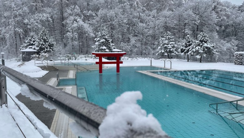 Ein verschneites Au&szlig;enbecken einer Therme mit roten asiatischen Elementen und schneebedeckten B&auml;umen im Hintergrund.