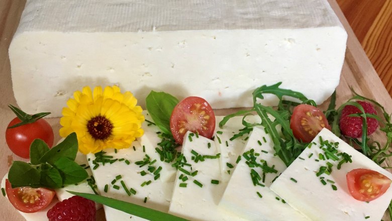 Ein Block Schafkäse in Scheiben geschnittenen, garniert mit Tomaten, Himbeeren, Rucola und einer gelben Blume auf einem Holzbrett.