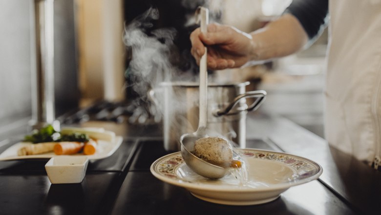 Eine Person schöpft Suppe mit einem Knödel aus einem Topf in einen Teller.