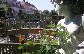Steinstatue neben einer Holzbrücke und roten Rosen im Vordergrund.