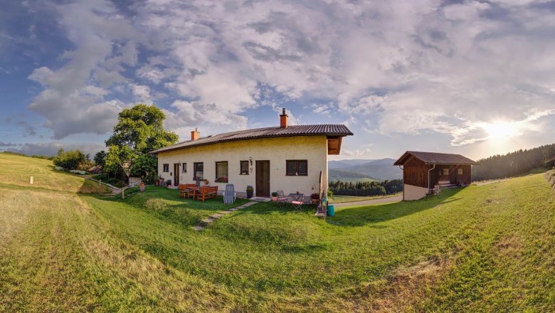 Panoramablick auf ein Haus auf einer Wiese mit B&auml;umen und Bergen im Hintergrund bei Sonnenuntergang.
