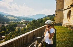 Eine Radfahrerin mit Helm steht vor einer alten Kirche, im Hintergrund der Ort Kirchberg am Wechsel und H&uuml;gel