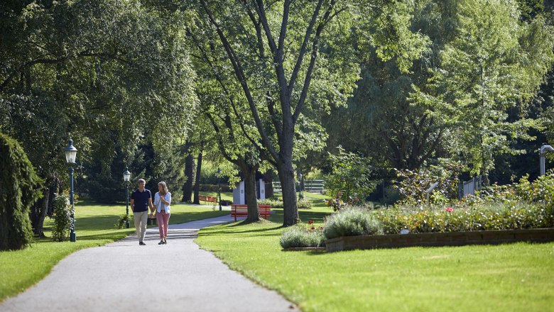 Ein Paar spaziert auf einem Weg im Kurpark Bad Sch&ouml;nau, umgeben von gr&uuml;nen B&auml;umen und gepflegten Blumenbeeten.