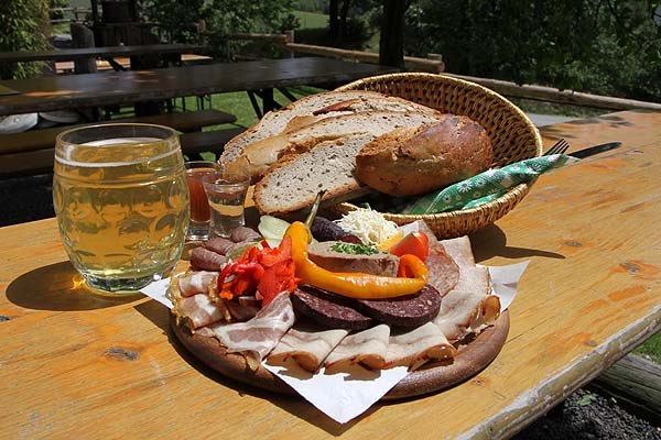 Brettljause mit Brot, Wurst, Speck und Getränken auf einem Holztisch im Freien.