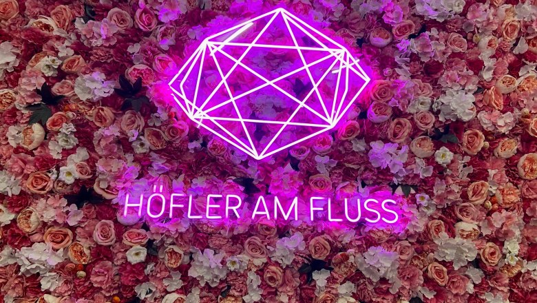 Pinker Neon-Schriftzug 'Höfler am Fluss' vor einer Wand aus rosa Blumen.