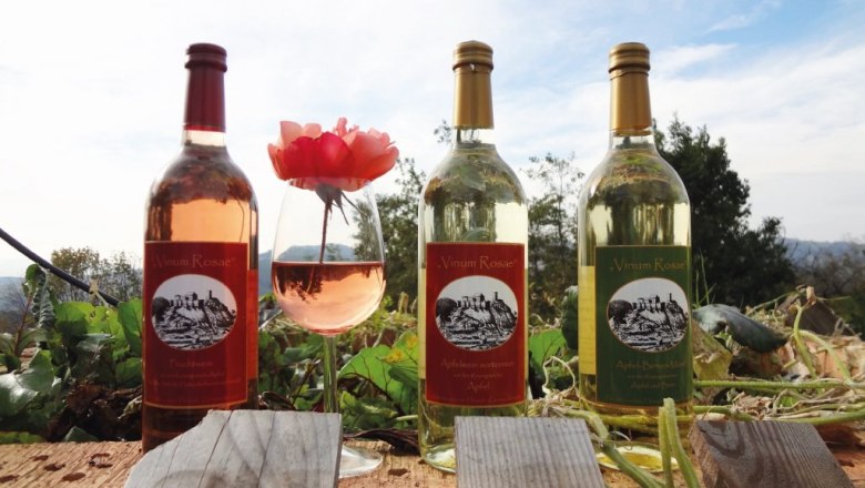 Drei Flaschen Apfelwein und ein Glas mit Apfelwein und einer Rose auf einem Holzbalken im Freien.