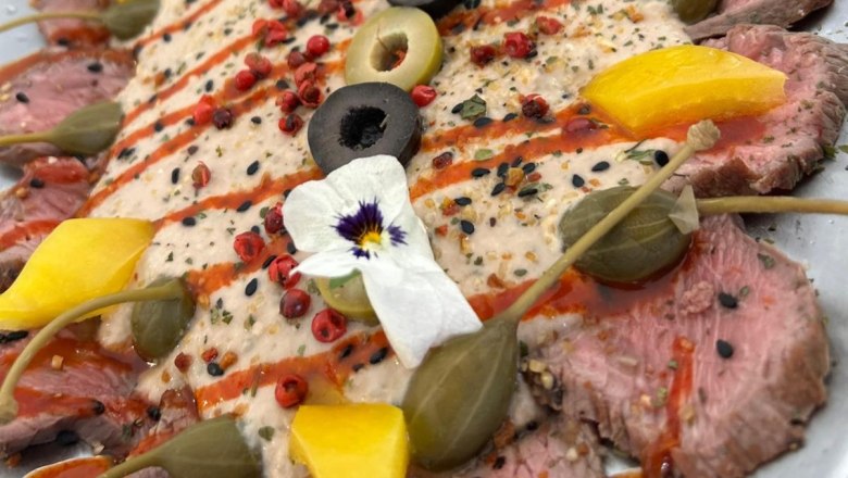 Nahaufnahme eines dekorativen Tellers mit rosa, feingeschnittenem Fleisch mit Kapern, Oliven und essbaren Blüten.