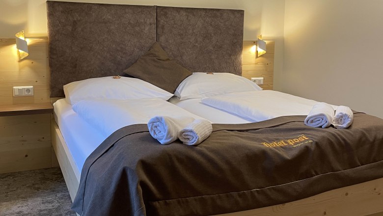 Ein gem&uuml;tliches Doppelzimmer mit gro&szlig;em Bett, dekoriert mit brauner Bettw&auml;sche und Handt&uuml;chern. An der Wand sind geometrische Muster.