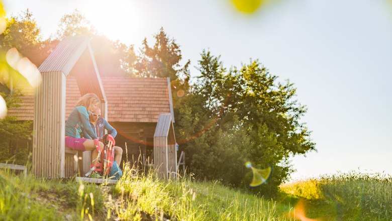 Zwei Personen sitzen in einer hölzernen Struktur in einer sonnigen Landschaft.