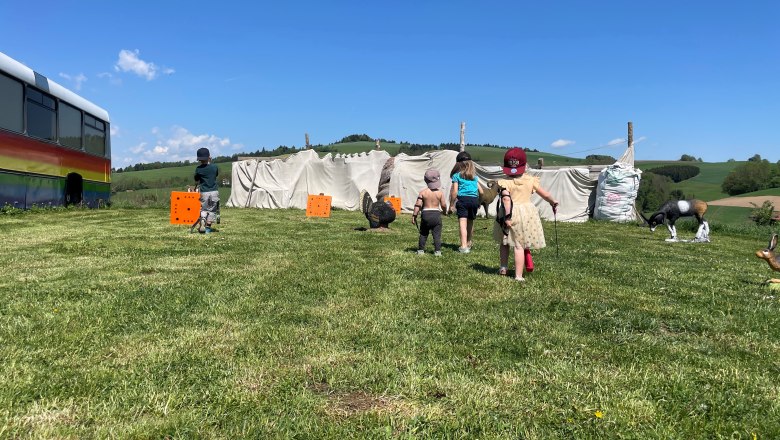 Kinder spielen auf einer Wiese mit Bogenschießzielen und einem bunten Bus im Hintergrund.