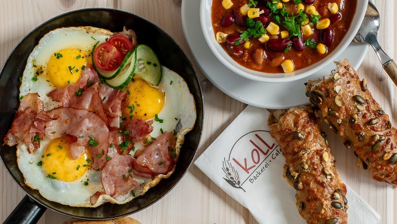 Verschiedene warme Snacks auf einem Tisch, darunter Spiegelei mit Speck, Chili con Carne, belegte Brote und ein Brötchen.
