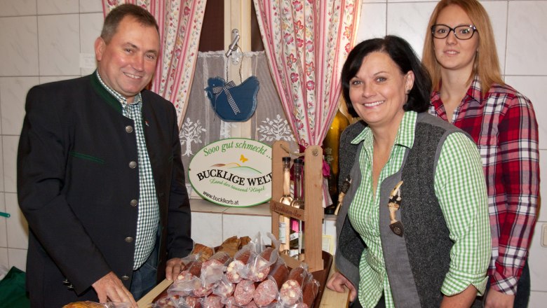 Drei Personen stehen in einem Hofladen vor einem Tisch mit Wurstwaren. Im Hintergrund ein Schild mit der Aufschrift 'Bucklige Welt'.