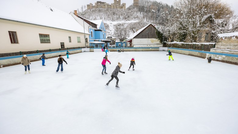 Menschen eislaufend auf einem Platz in Kirchschlag, im Hintergrund auf einem H&uuml;gel eine Burgruine.
