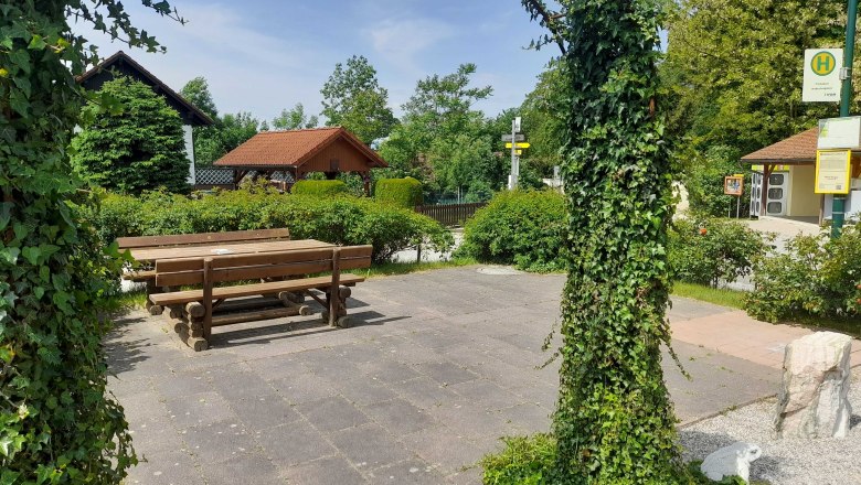 Ein ruhiger Platz mit Holzbänken und Tischen, umgeben von grünen Pflanzen und einem mit Efeu bewachsenen Torbogen.