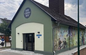 Ein kleines, grünes Gebäude mit der Aufschrift 'Dorfladen Box' und einem gemalten, ländlichen Wandbild.